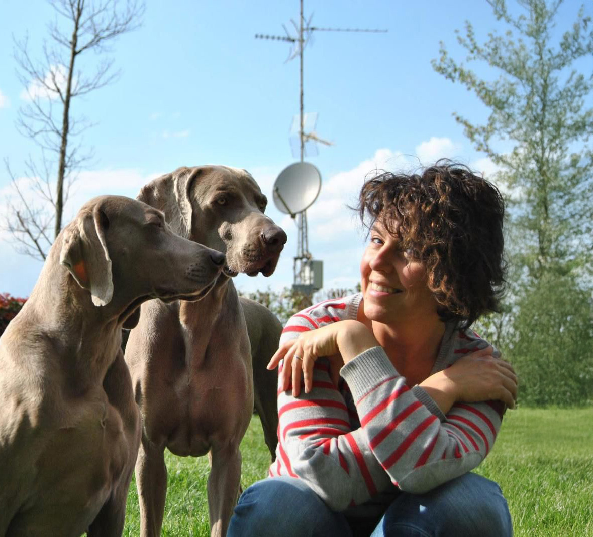 Woman with glasses sitting on grass outdoors with two light brown dogs resting paws on her legs.