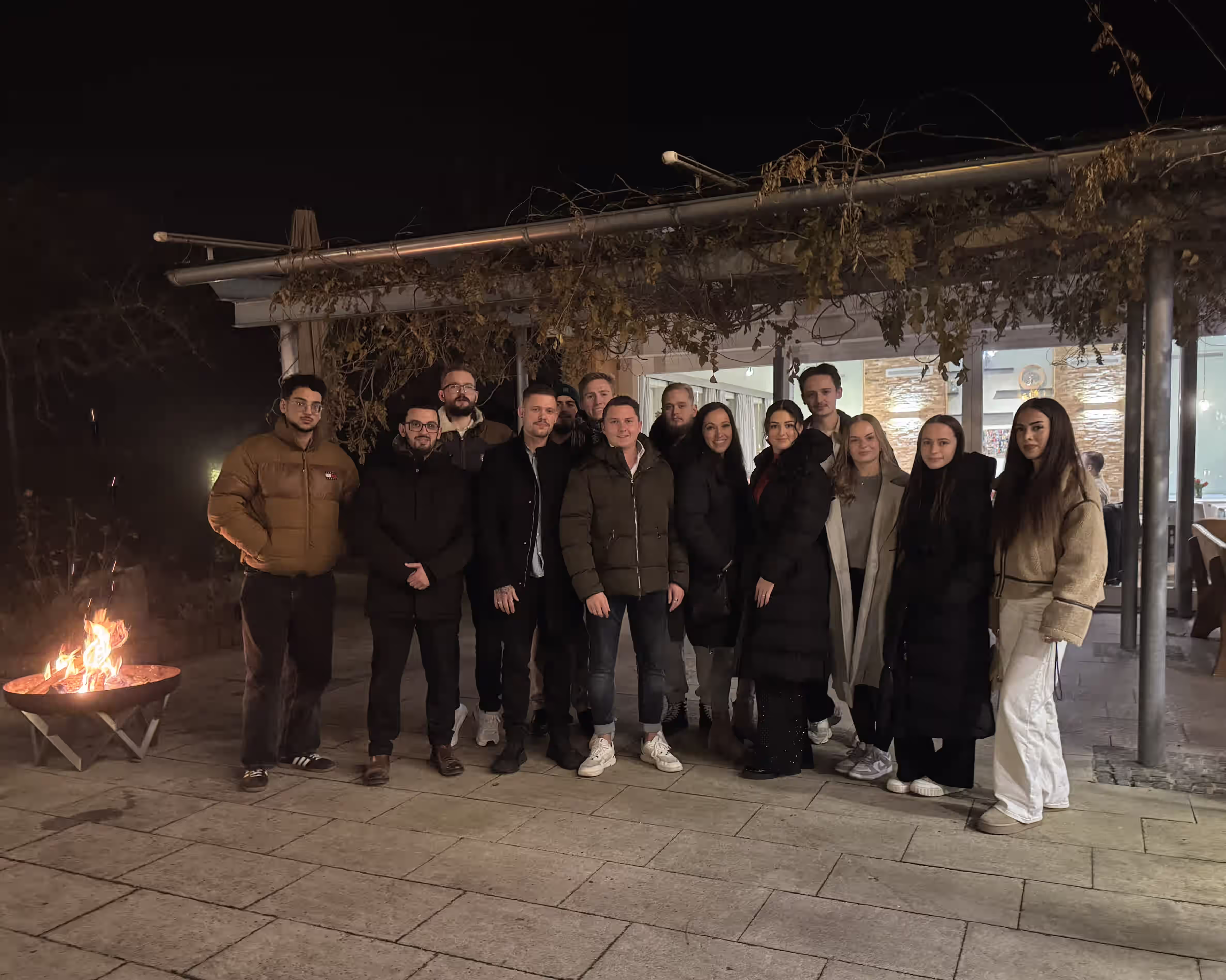 Gruppe von 14 Personen steht bei Nacht draußen neben einem Feuerkorb vor einem beleuchteten Gebäude.