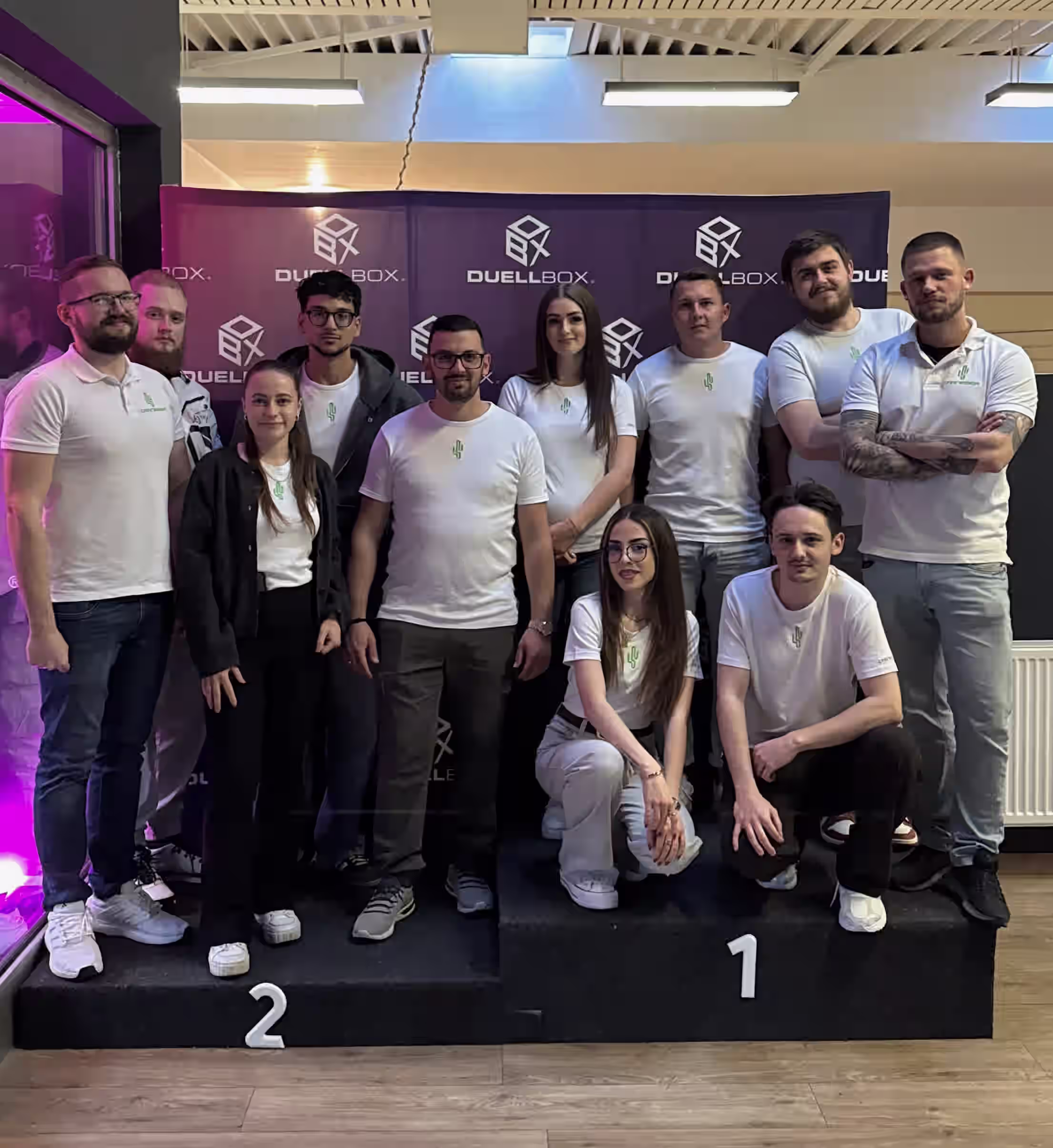 Gruppenfoto von elf Personen, die auf Podesten mit den Nummern 1 und 2 vor einer Duelbox-Wand posieren.