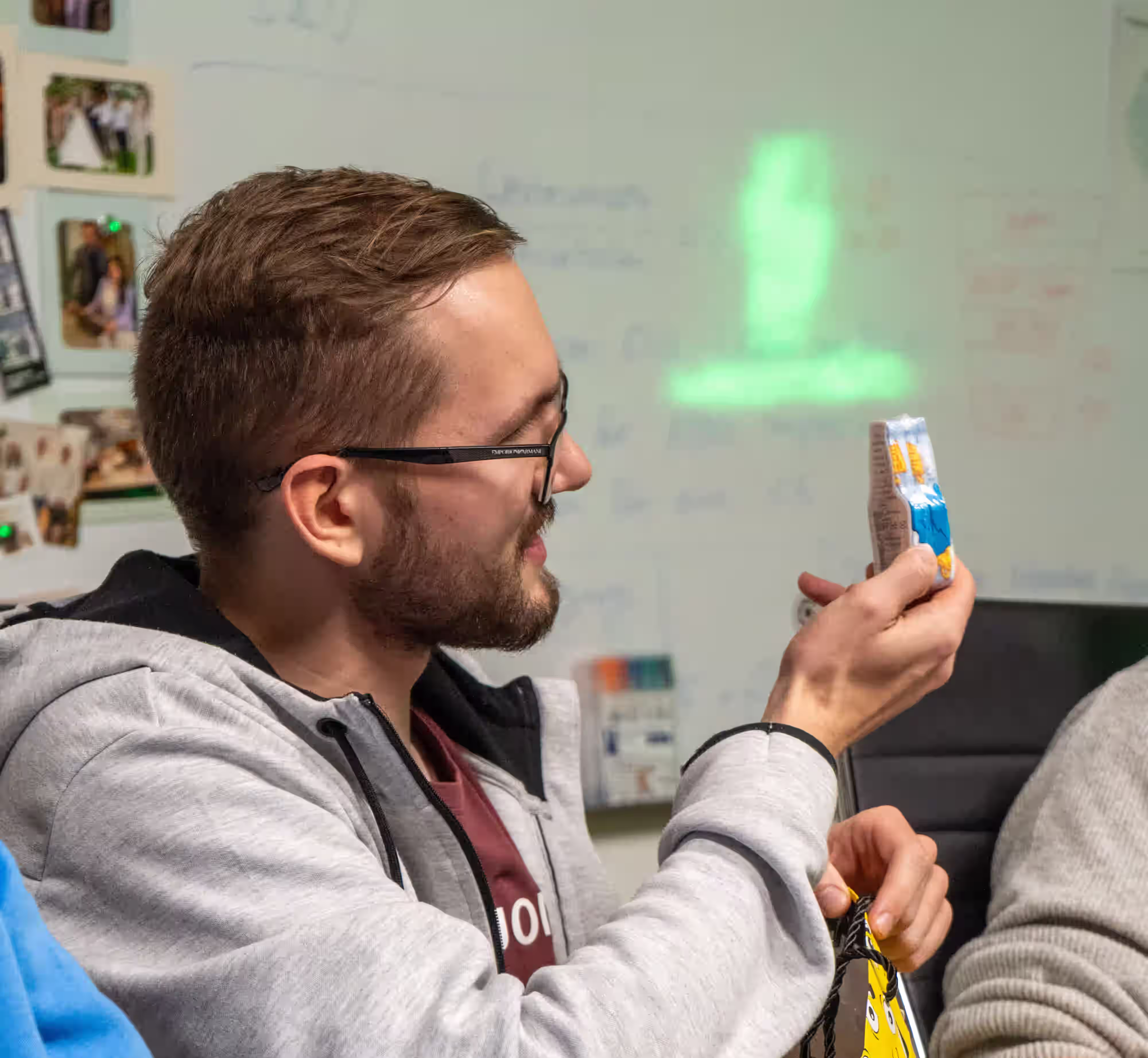 Mann mit Brille hält lächelnd eine Snackverpackung in einem Büro mit Whiteboard im Hintergrund.