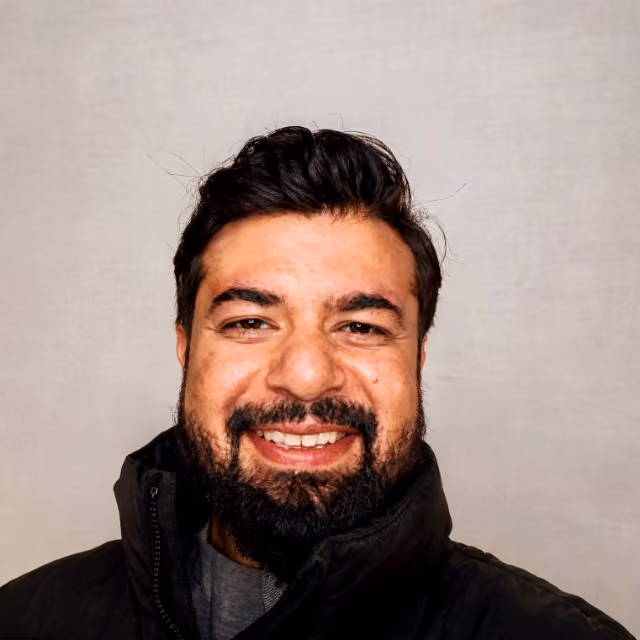 Smiling man with dark hair and beard wearing a black jacket against a plain light background.