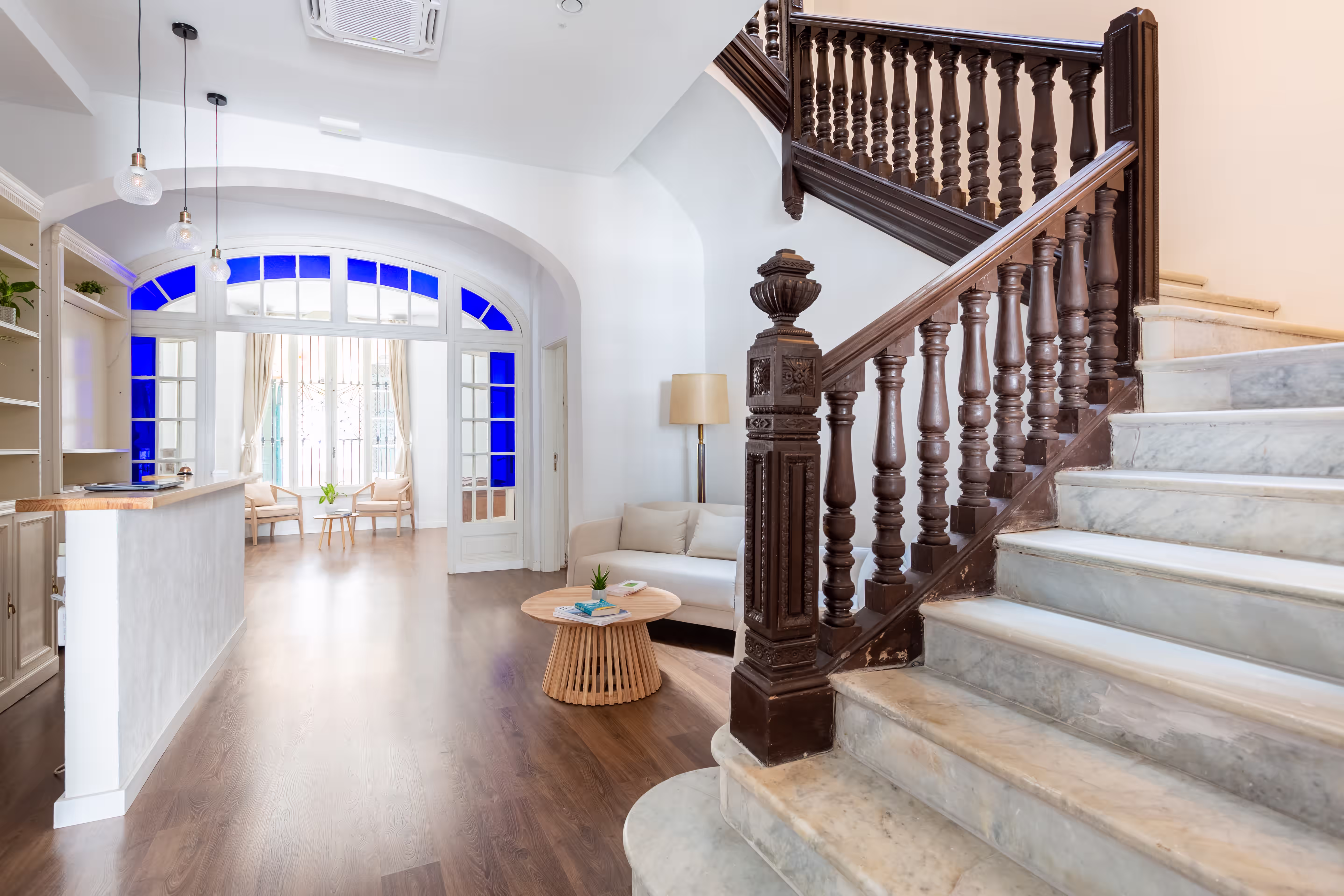 Bright lobby with wooden floor, marble staircase, white sofa, round table, and blue stained glass windows.