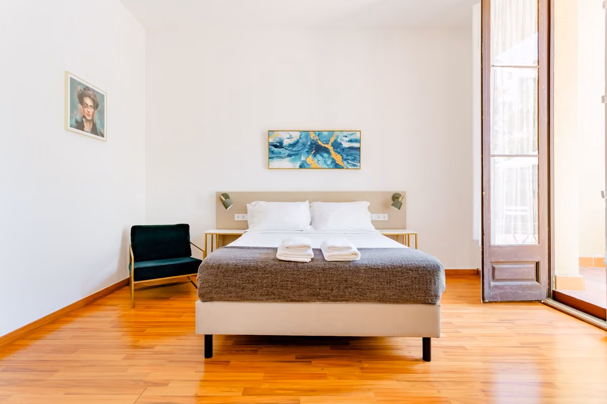 Minimalist bedroom with a double bed, two bedside tables, wall lamps, a beige armchair, and a large painting.