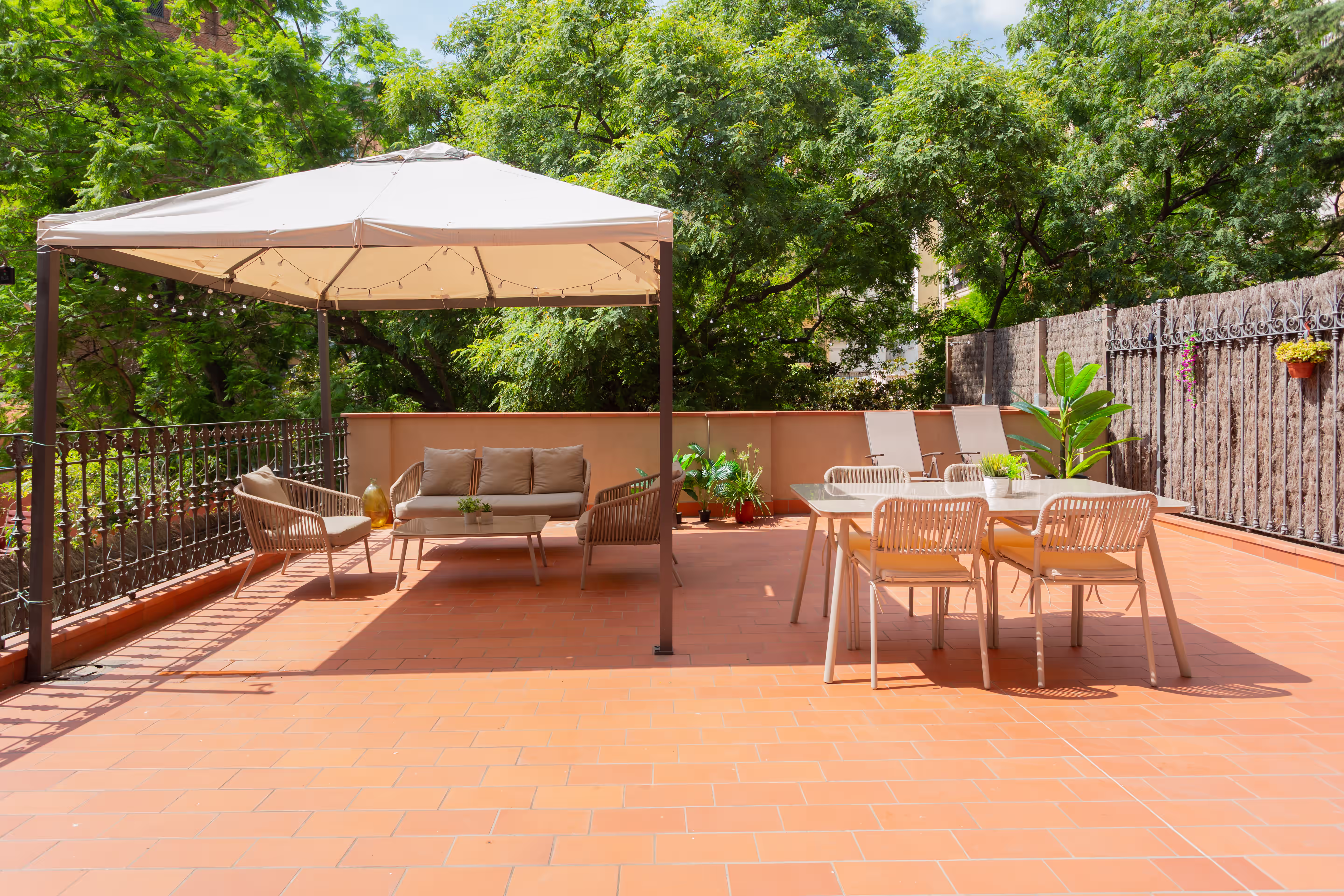 Sunlit outdoor patio with wicker seating under canopy, dining table with chairs, and two lounge chairs.