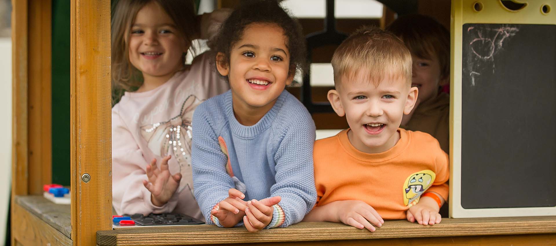 Three smiling children looking out of a wooden play house
