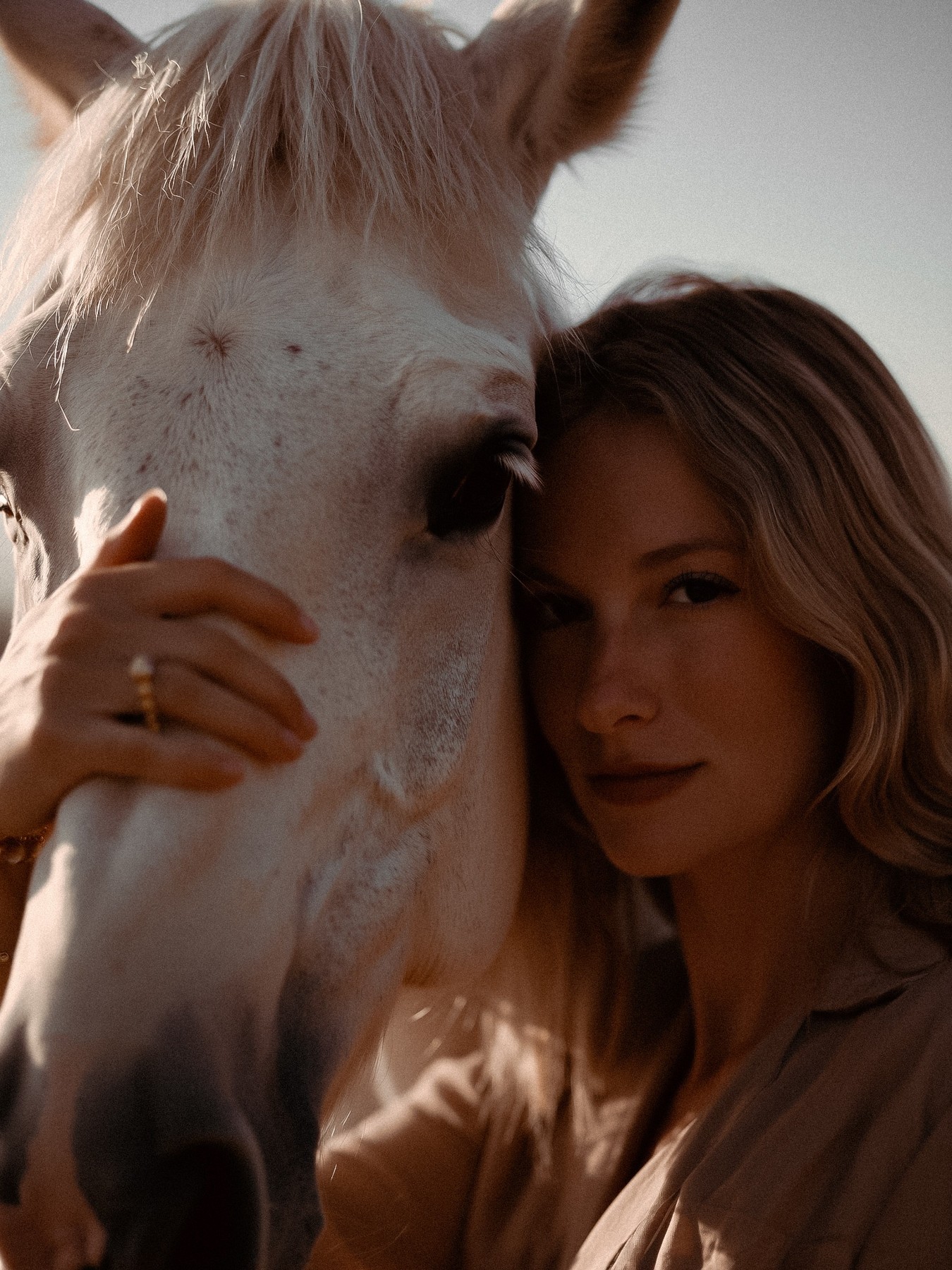 A woman rests beside a white horse in soft light.