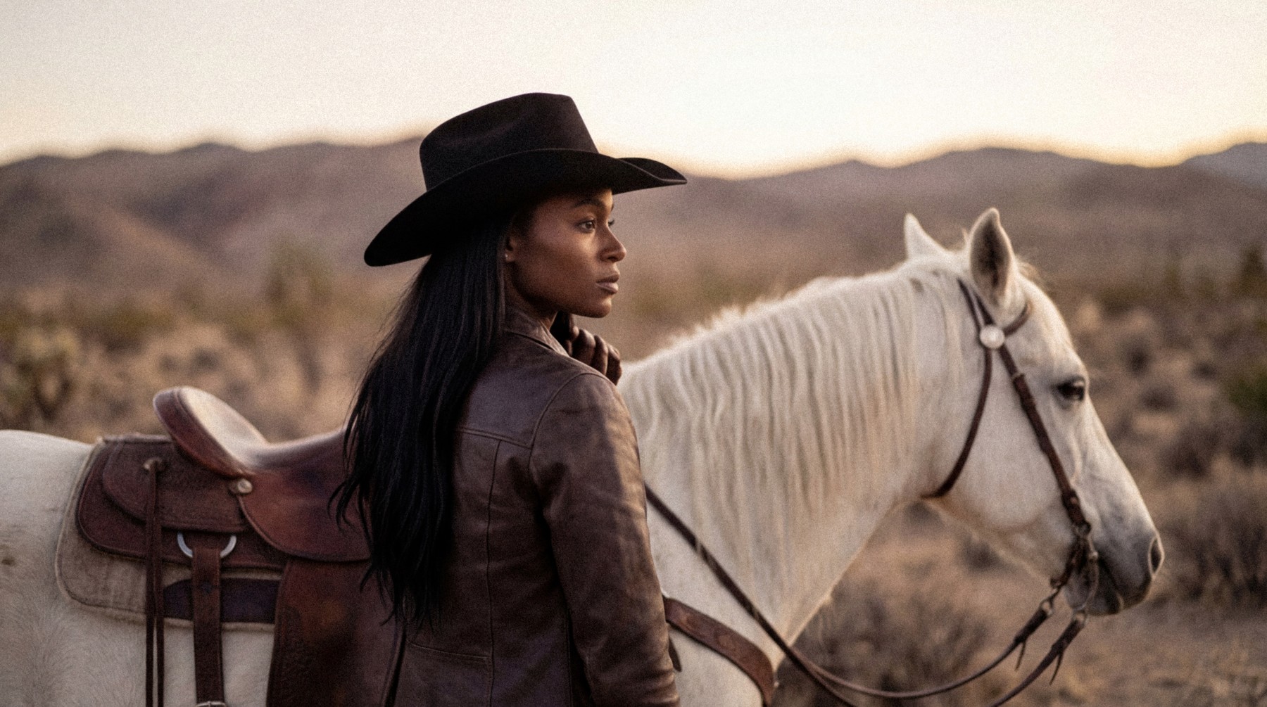 A woman in a cowboy hat beside a white horse in the desert.