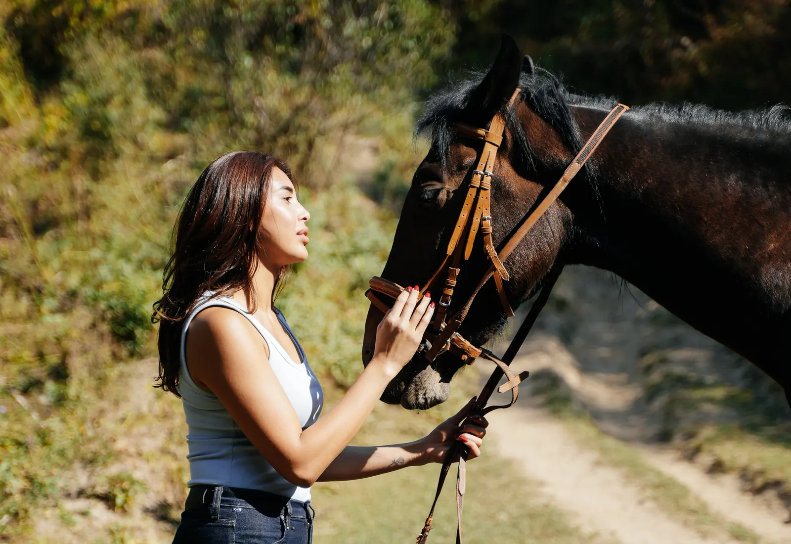 A woman standing beside a dark horse, gently touching its face on a leafy path.