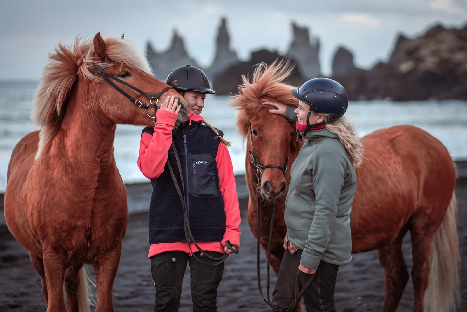 Two helmeted riders with chestnut horses on the black sand beach.