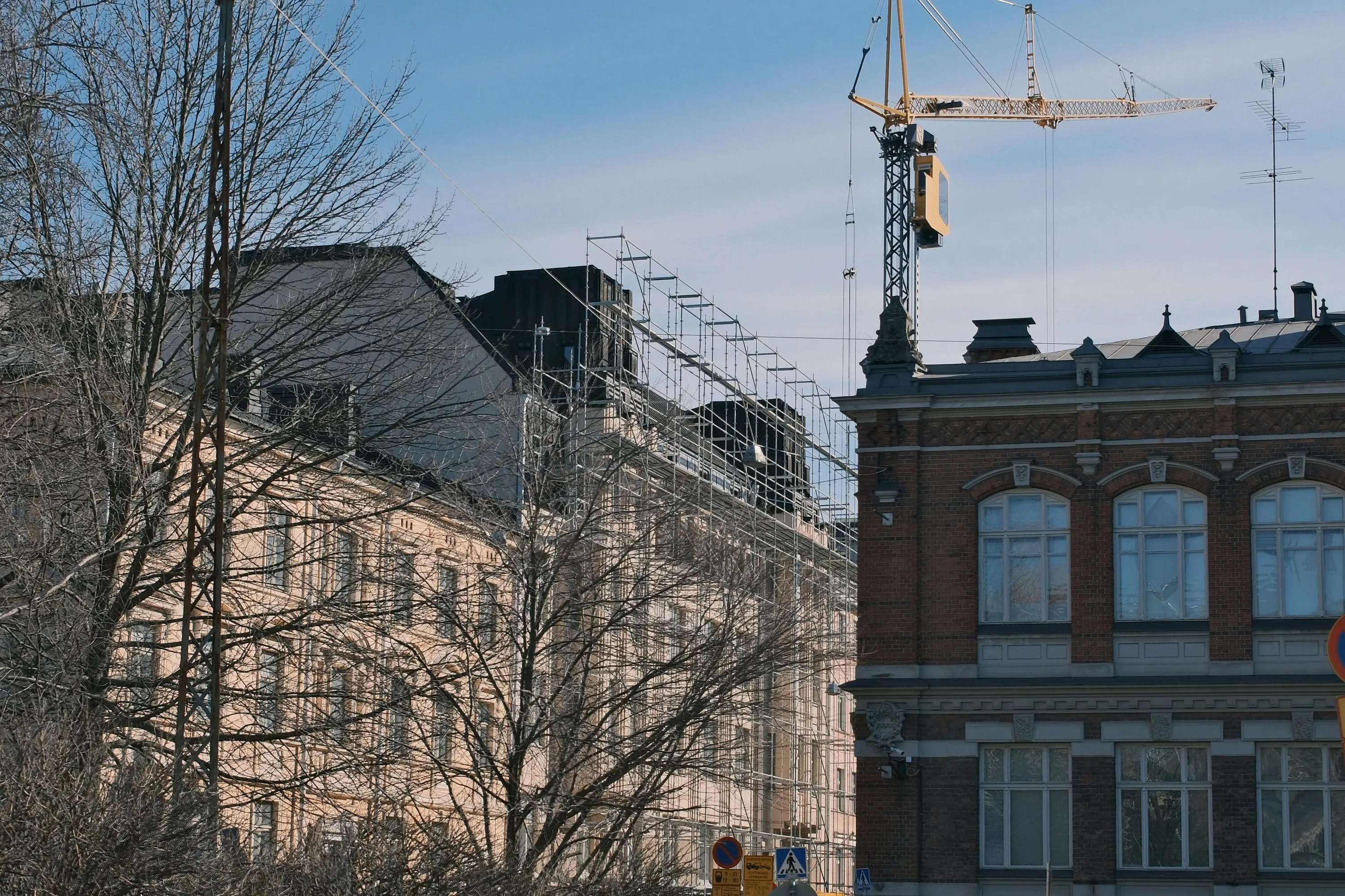 Aurinkoinen kuva kaupunkimaisemasta, jossa on rakennuksia, puita ja rakennustyömaa. Vasemmalla on tiheäoksaisia, lehdettömiä puita. Niiden takana näkyy vaaleanpunertava rakennus, jonka julkisivua peittävät rakennustelineet. Oikealla etualalla on punatiilinen rakennus, jossa on kaarevia ikkunoita ja yksityiskohtaisia muotoiltuja elementtejä katonrajassa ja ikkunoiden yläpuolella. Tämän rakennuksen katolla on antenni. Taivaalla, rakennusten yläpuolella, kohoaa suuri keltainen rakennusnostokurki. Taivas on kirkkaan sininen.