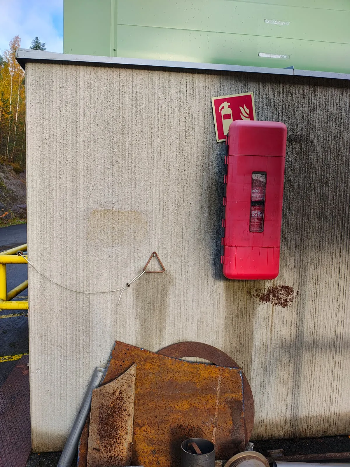 Kuva vaaleasta betoniseinästä, jossa on punainen palosammutinkyltti ja -kaappi oikealla puolella. Seinällä roikkuu ruosteinen metallikolmio ohuella vaijerilla. Seinän alaosassa on kasa ruosteisia metallilevyjä ja putkia. Taustalla näkyy vihreä rakennuksen yläosa, sininen taivas ja syksyisiä puita sekä keltainen metalliaita vasemmalla. Päivänvalo valaisee kohteen.