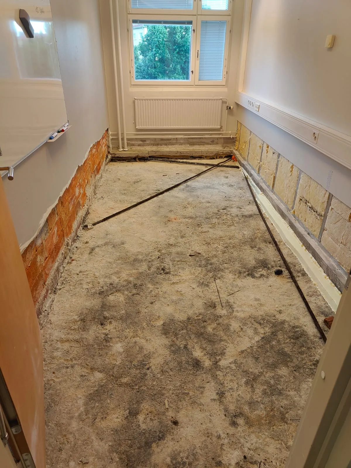 Remontoitava huone, jossa on paljas betonilattia. Vasemmalla seinällä näkyy punatiiliä ja yläosassa valkoinen taulu. Oikealla seinällä on esillä keltaista eristettä ja pistorasioita. Takaseinällä on ikkuna, josta näkyy puita, ja valkoinen patteri. Lattialla on metallitankoja ja likaa. Huone on valaistu luonnonvalolla.