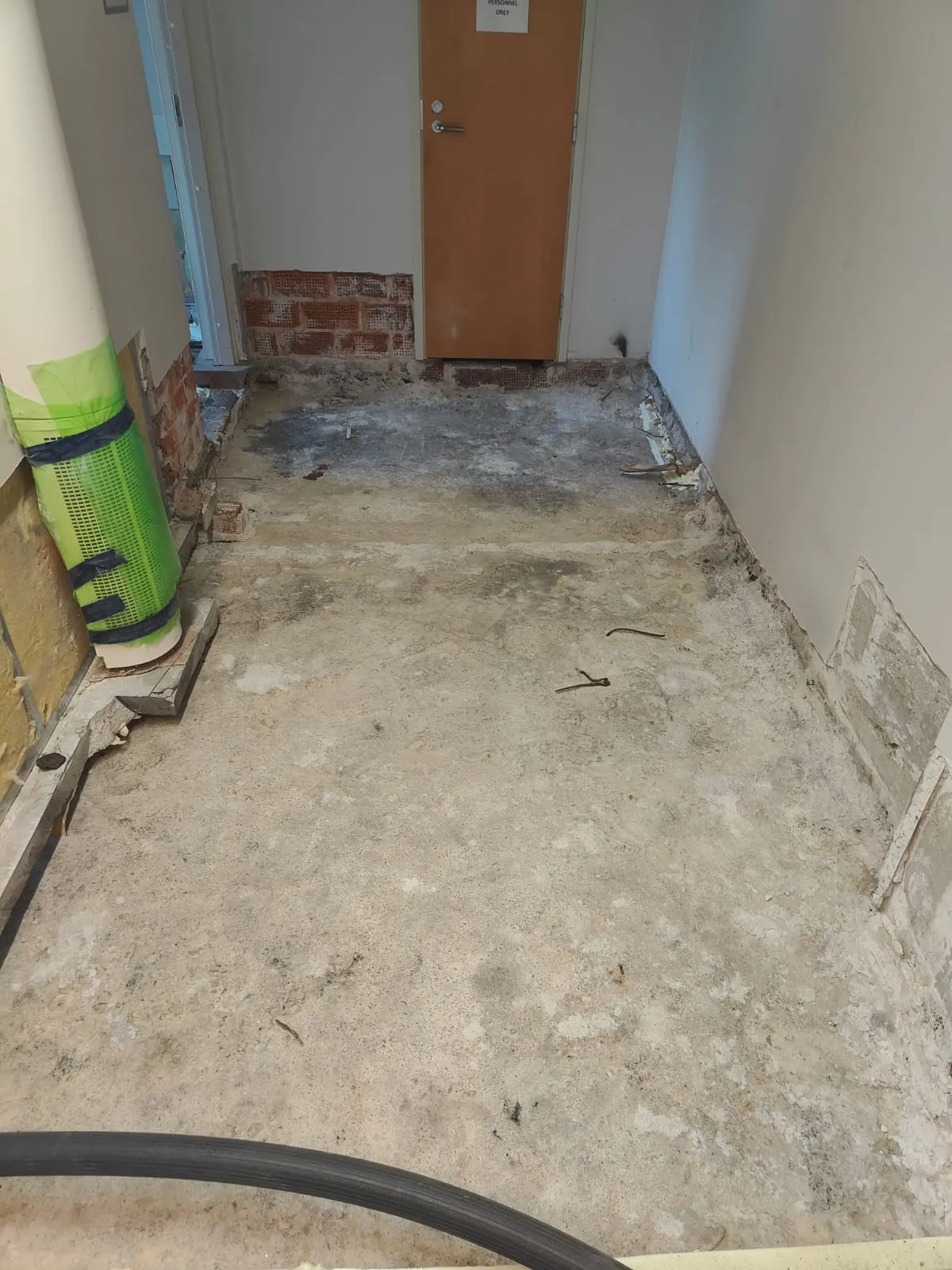 Remontissa oleva sisätila, jossa paljas, pölyinen ja epätasainen betonilattia. Vasemmalla on osittain tiiliverhoiltu seinä ja suuri valkoinen putki, jossa on vihreä verkko. Takaosassa on ruskea puuovi, jonka yläpuolella on kyltti 'PERSONNEL ONLY', ja sen ympärillä näkyy tiiliseinää ja valkoiseksi maalattua seinää. Oikea seinä on valkoinen, ja sen alaosassa on vaurioita. Lattialla on kaksi taivutettua metallikappaletta ja kuvan alareunassa musta letku. Valaistus on tasainen.