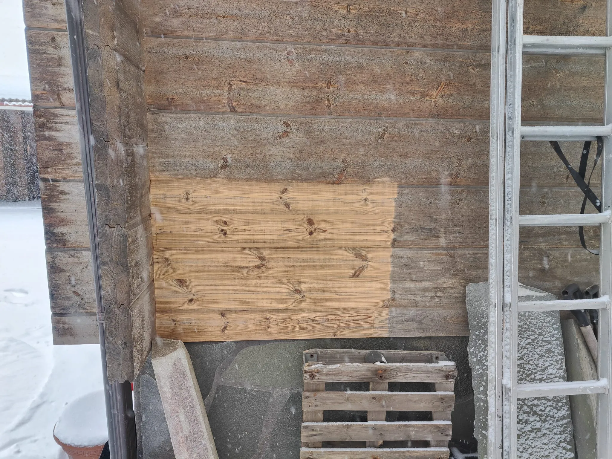 Ulkokuva puuseinästä lumisateessa. Seinän yläosa ja oikea puoli ovat harmaantunutta puuta. Keskellä on vaaleampi, hiottu puualue, joka paljastaa kellertävänruskean pinnan. Vasemmalla näkyy rakennuksen nurkka ja tumma sadevesiputki. Maassa on lunta. Seinän edessä on lumipeitteinen puinen kuormalava, vaaleita rakennuspalikoita ja harmaita laattoja. Oikealla nojaa seinää vasten hopeanvärinen, lumipeitteinen tikkaat.