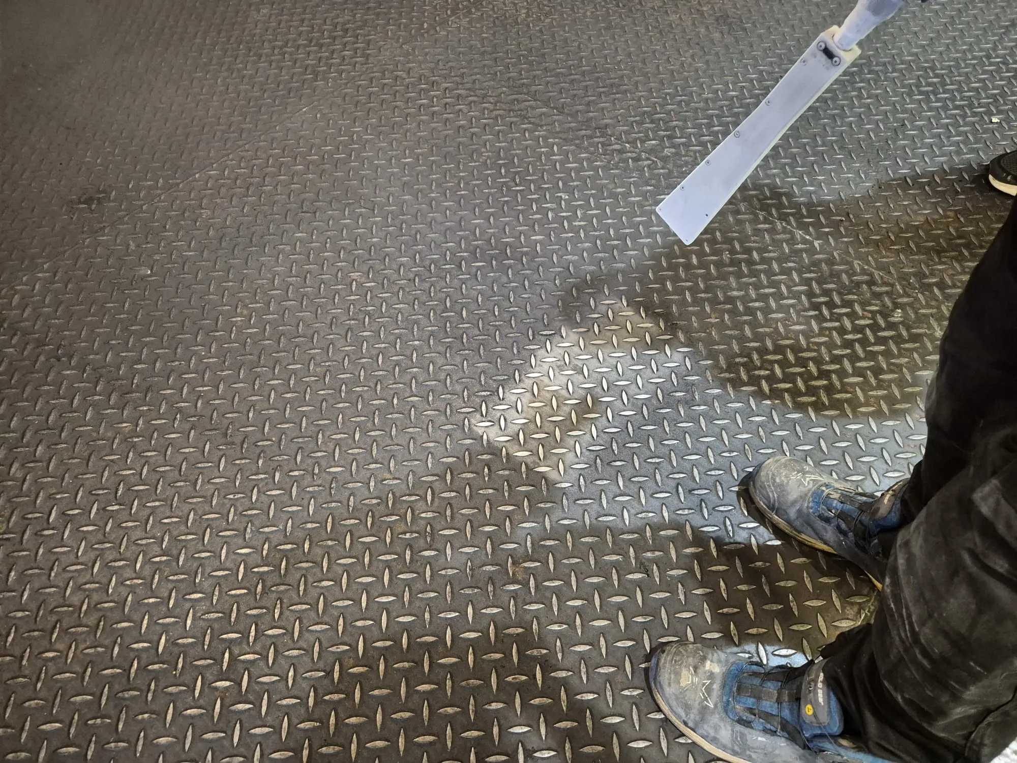 Tummanharmaa metallilattia, jossa on ruutukuvio. Oikeassa alakulmassa näkyy henkilön jalat likaisissa sinisissä työkengissä ja tummissa housuissa. Oikeassa yläkulmassa on vaalea, litteä esine, joka heijastaa kirkasta valoa lattialle ja luo varjon. Lattian pinta on kulunut ja heijastaa valoa epätasaisesti.