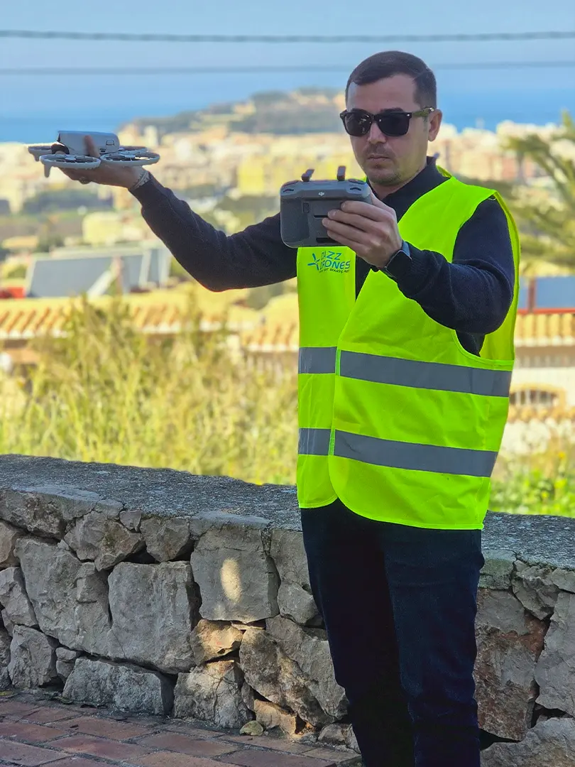 Dazz Drones pilot wearing a high-visibility vest, holding a drone and controller with a coastal cityscape in the background.