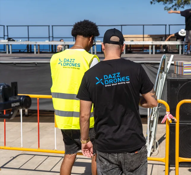 Two Dazz Drones team members walking through an outdoor work area, wearing branded shirts and a high-visibility vest.