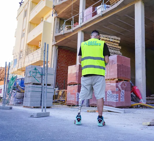 Dazz Drones pilot wearing a high-visibility vest at a construction site, with building materials and a partially constructed building in the background.