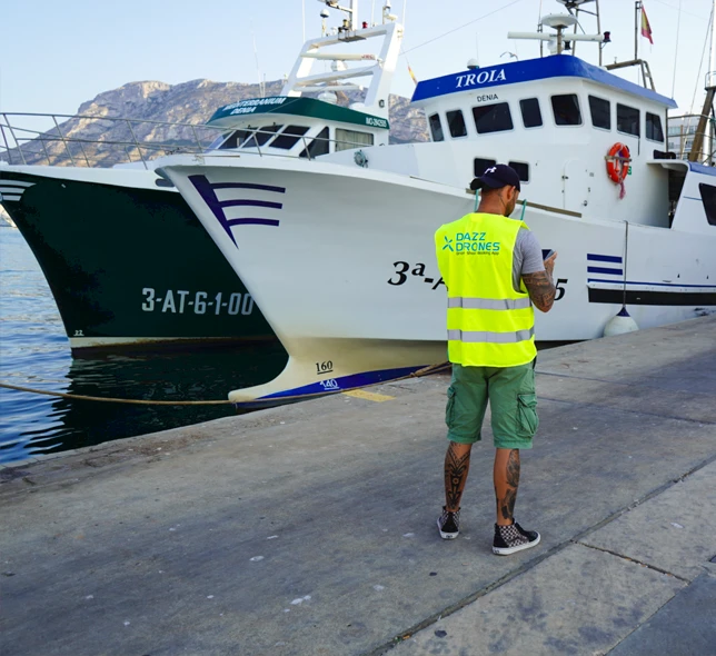Dazz Drones pilot wearing a high-visibility vest at a marina, standing beside two boats with mountains in the background.