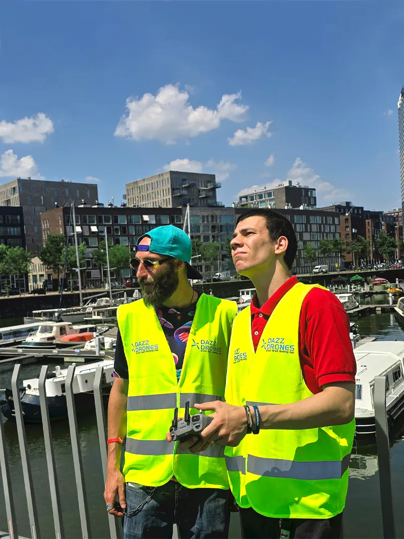 Two Dazz Drones team members wearing high-visibility vests at a marina, with a drone controller and boats in the background.