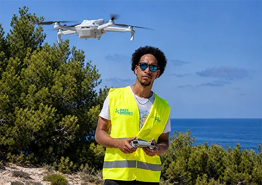 A person wearing a high-visibility vest stands outdoors holding a drone controller, with a white drone flying overhead and the ocean visible in the background.