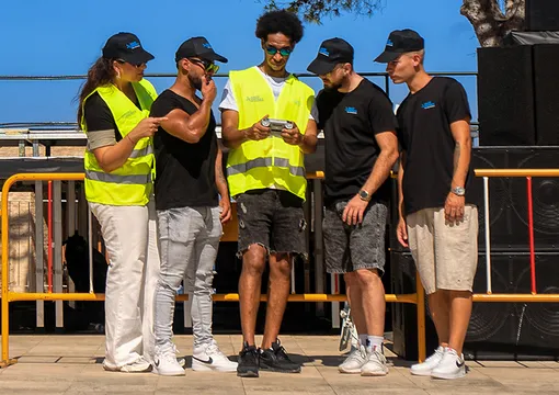 A group of five people, including event staff in high-visibility vests, standing together outdoors near a waterfront and looking at a smartphone.