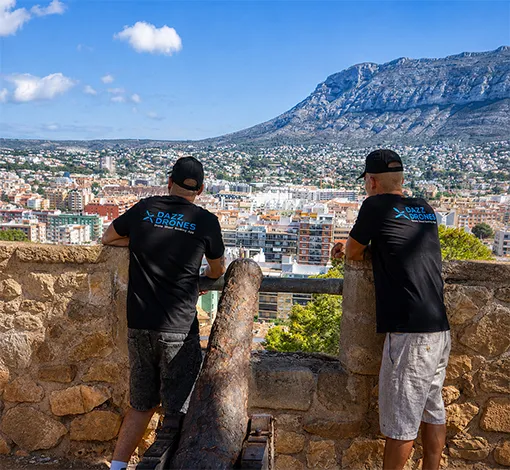 wo people in black shirts stand beside an old stone cannon overlooking a coastal city, with a large mountain rising in the background.