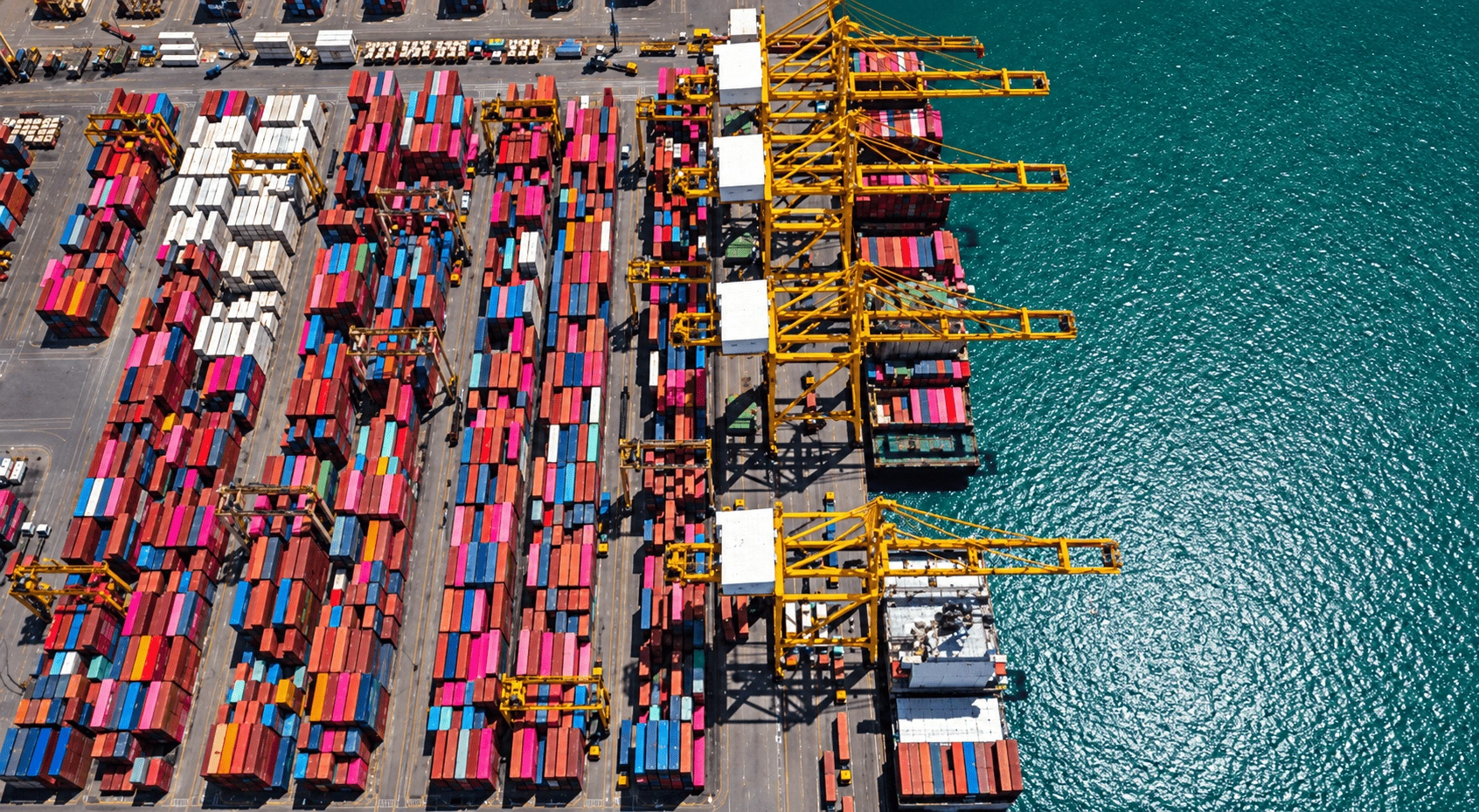Aerial drone view of a large industrial shipyard with cranes, construction equipment, storage areas, and vessels along the waterfront.