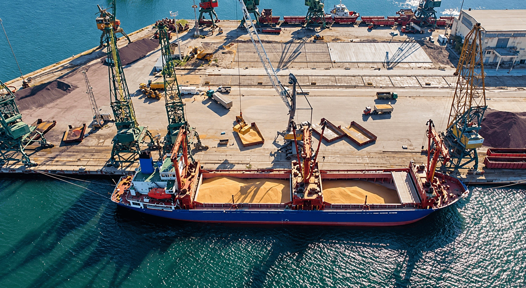 Aerial drone view of an industrial port with cargo ships, cranes, storage facilities, and heavy equipment along the waterfront.