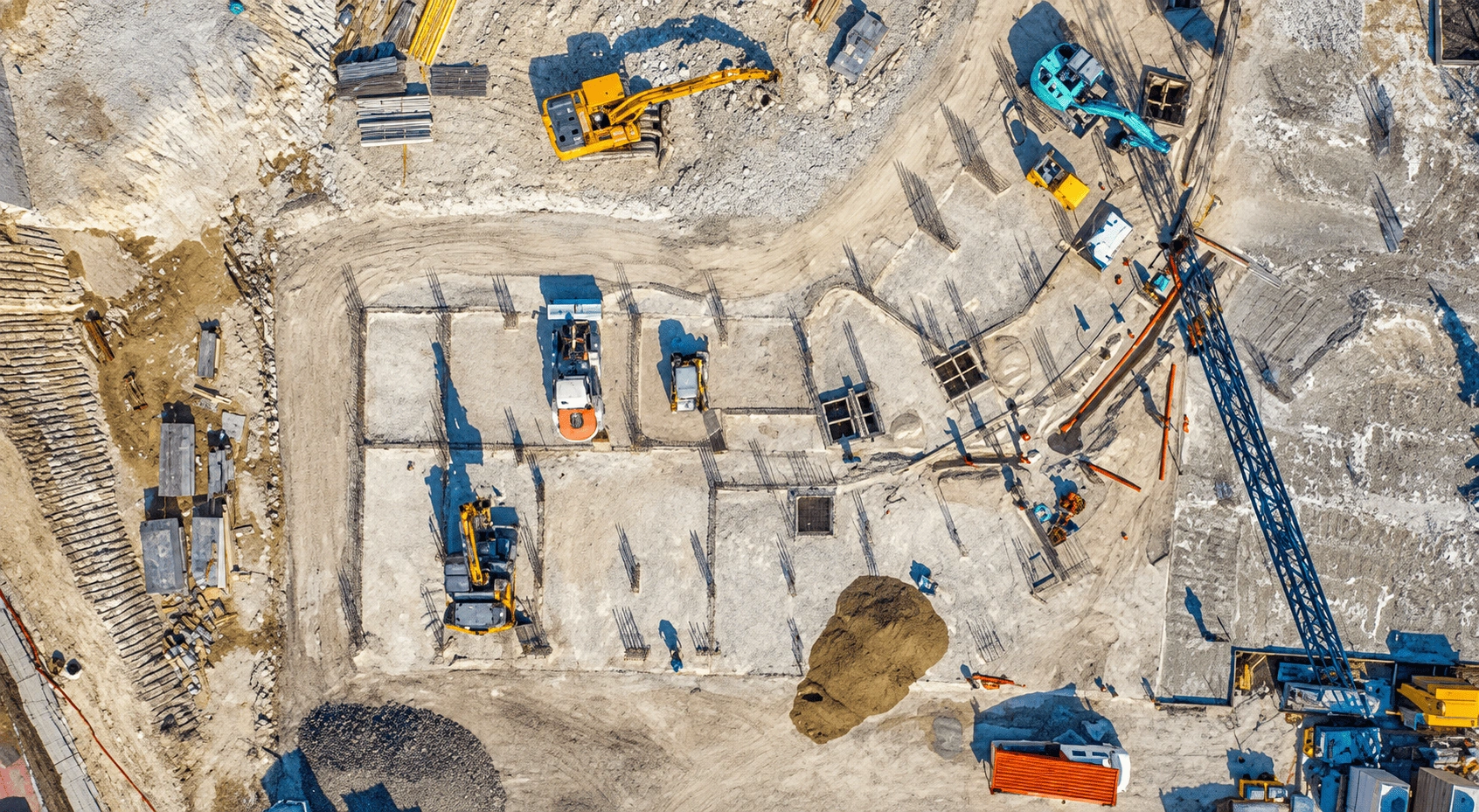 Aerial drone view of a construction site along a turquoise waterway, with heavy equipment and a large structure under construction.
