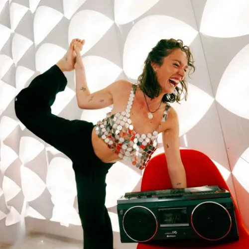 Founder and instructor Morgane Gillard, woman in a shiny silver top doing a yoga pose with one leg raised behind her while smiling next to a red chair and boombox.