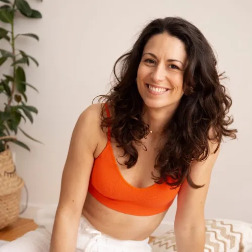 Aurore Martano, CAMOH's instructor, smiling woman with long curly hair wearing an orange sports bra sitting indoors near a plant.