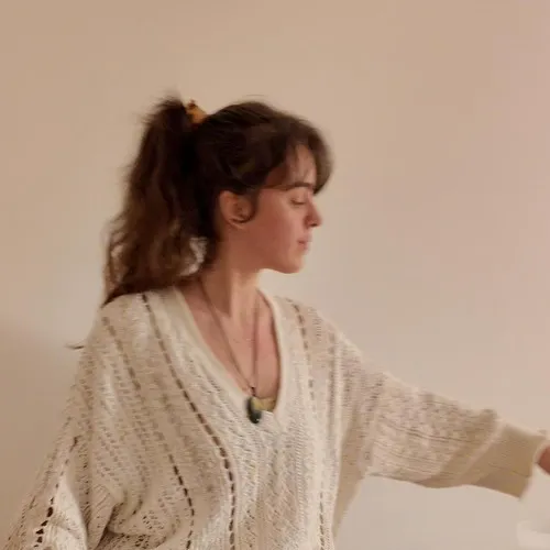 Claire, Sound Therapist & Energy Practitioner, Woman with brown hair in a ponytail wearing a cream knit sweater standing against a plain beige wall.