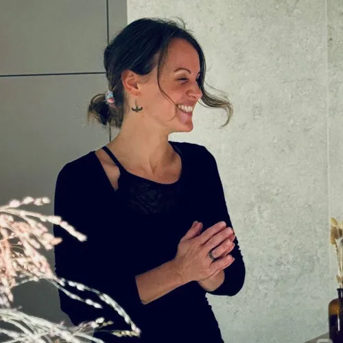 Sarah, Facialist, Body Therapist & Energy Practitioner as a Smiling woman with dark hair in a low bun wearing a black top, standing indoors with hands clasped.