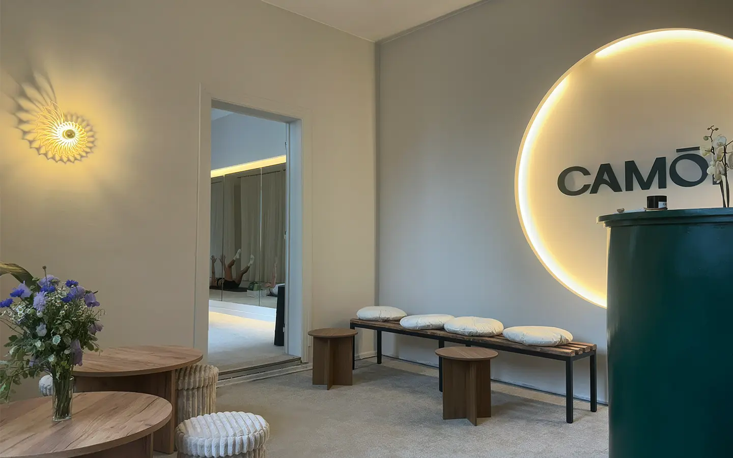 Minimalist waiting area with wooden tables, cushioned bench, round stools, flowers, and illuminated CAMÓH sign.