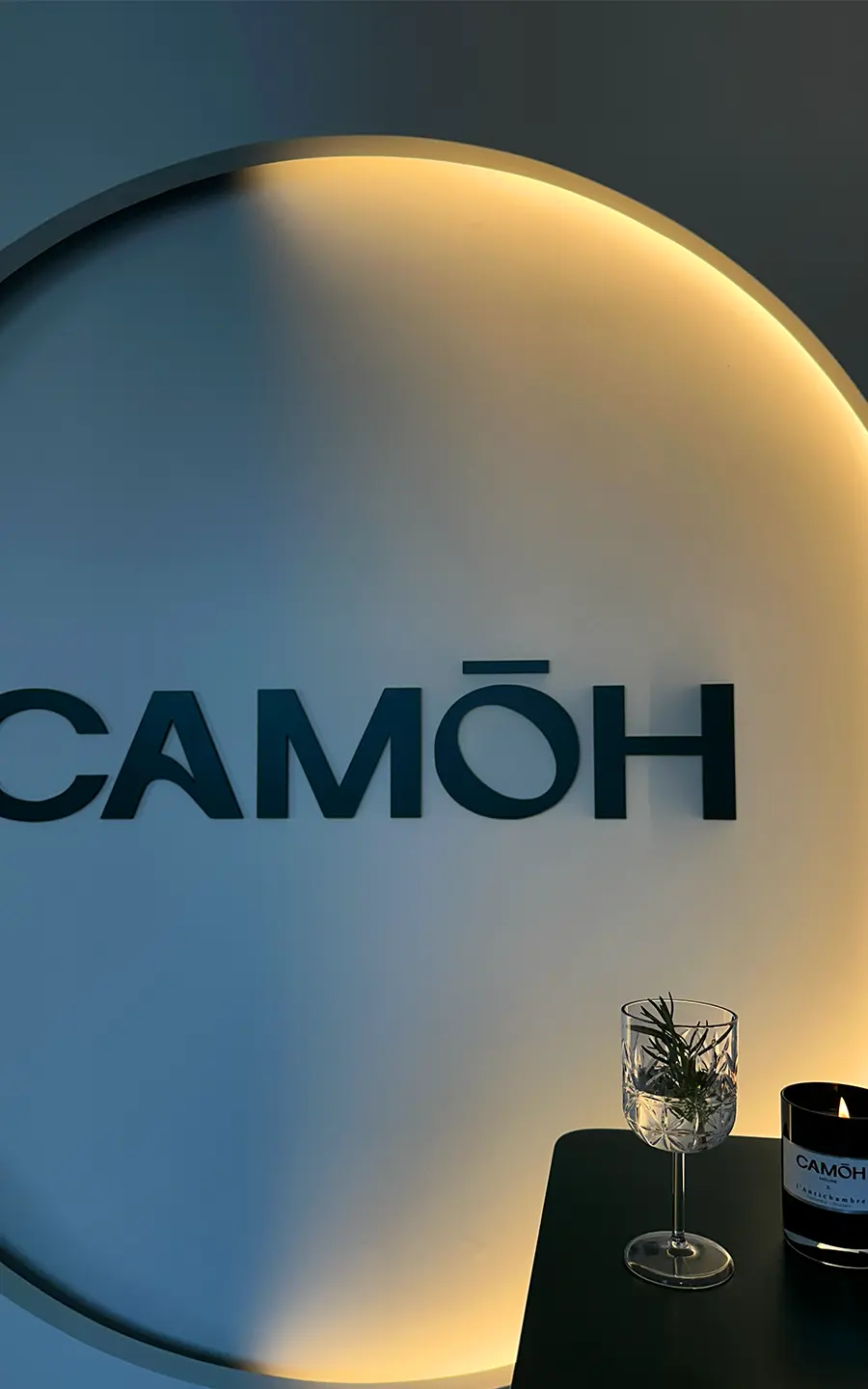 Dimly lit wall with CAMŌH sign, a glass with herbs, and a candle on a table.