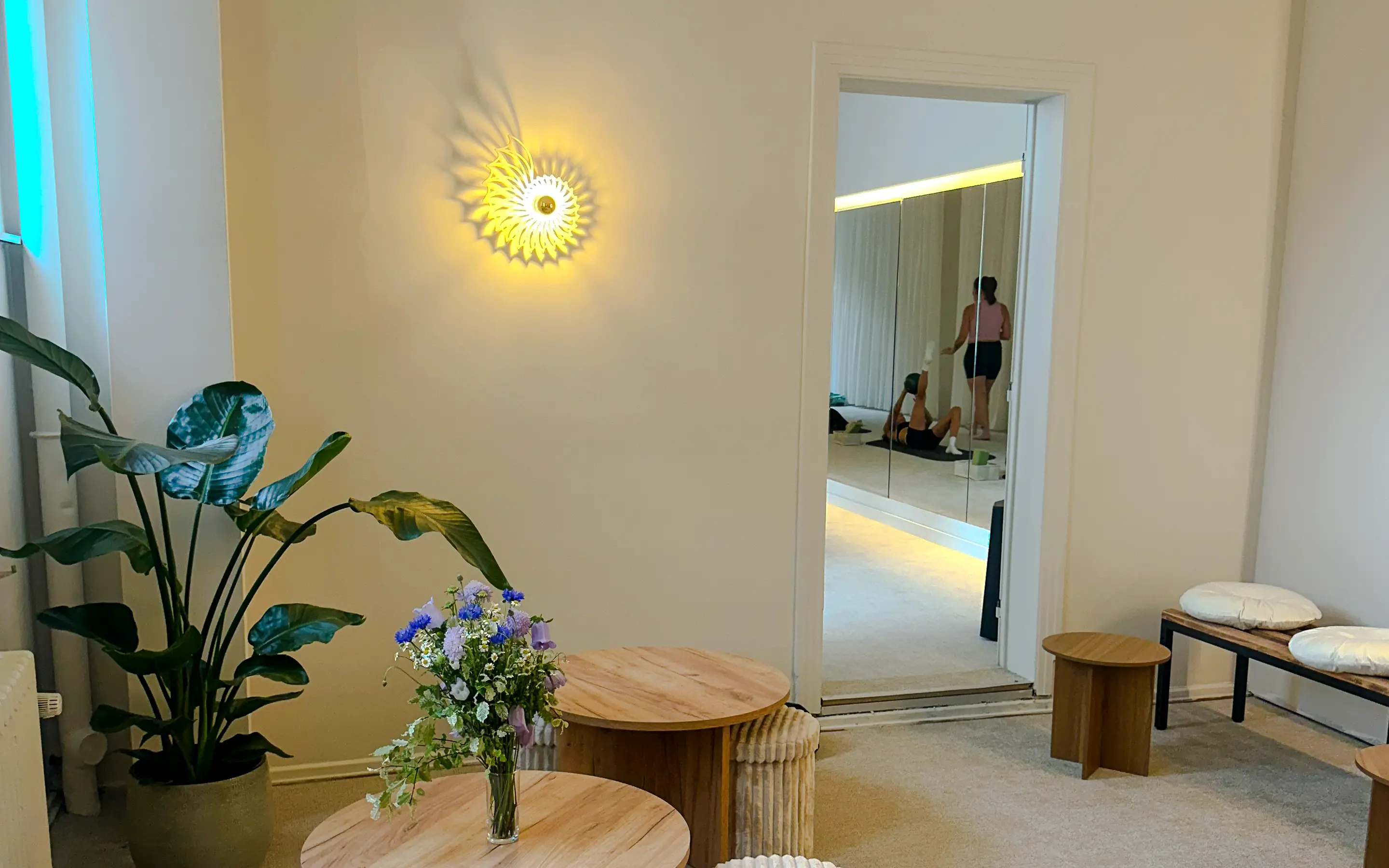 Cozy room with a plant, flower vase on wooden tables, wall light, and pilates class visible through doorway.