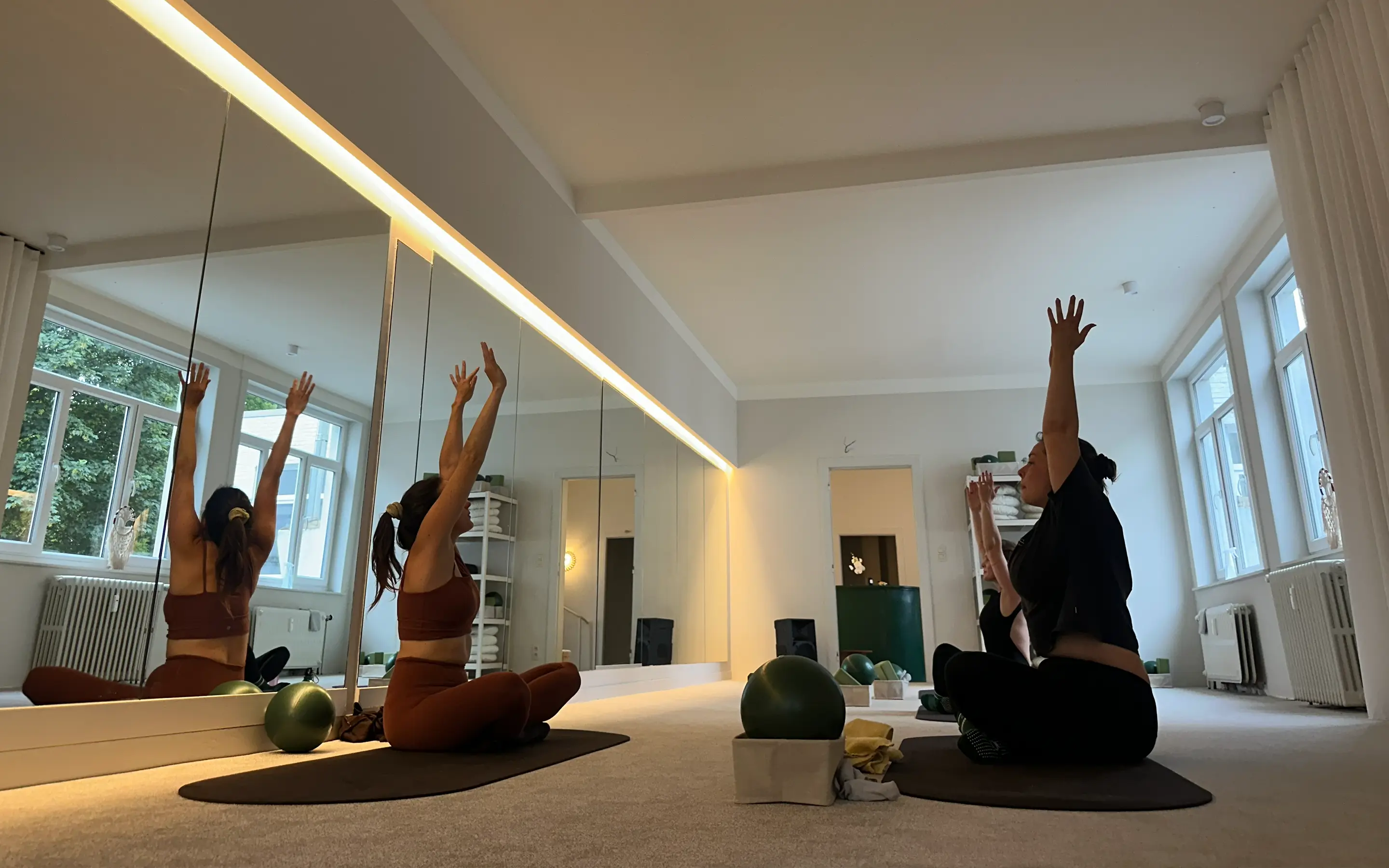 Three women sitting cross-legged on mats in a studio raising their arms during a pilates session.