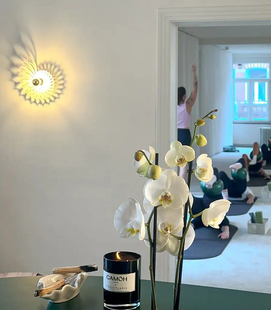 White orchid, lit candle, and smudge sticks on table with pilates class seen through doorway.
