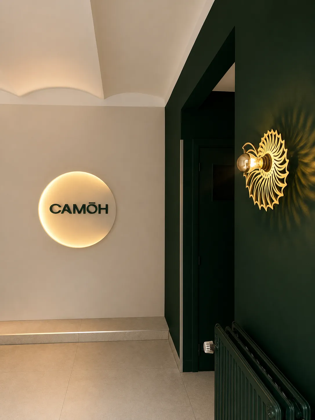 Interior with a beige wall, glowing circular sign with CAMÒH text, dark green wall, and golden wall light.