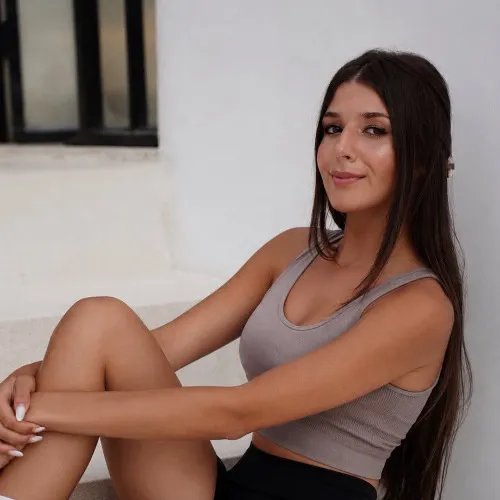 Vanessa, reformer teacher at CAMOH, with long dark hair sitting relaxed with arms wrapped around knees against a white wall.