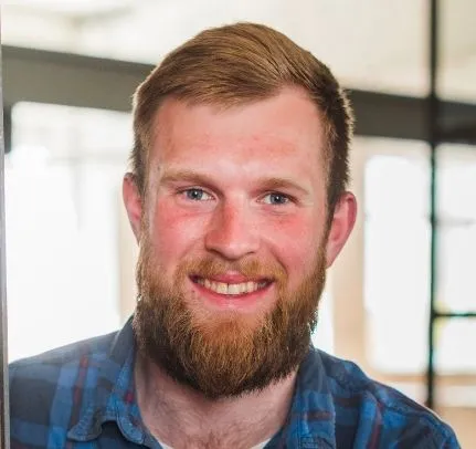 Smiling young man with red hair and beard wearing a blue plaid shirt indoors.