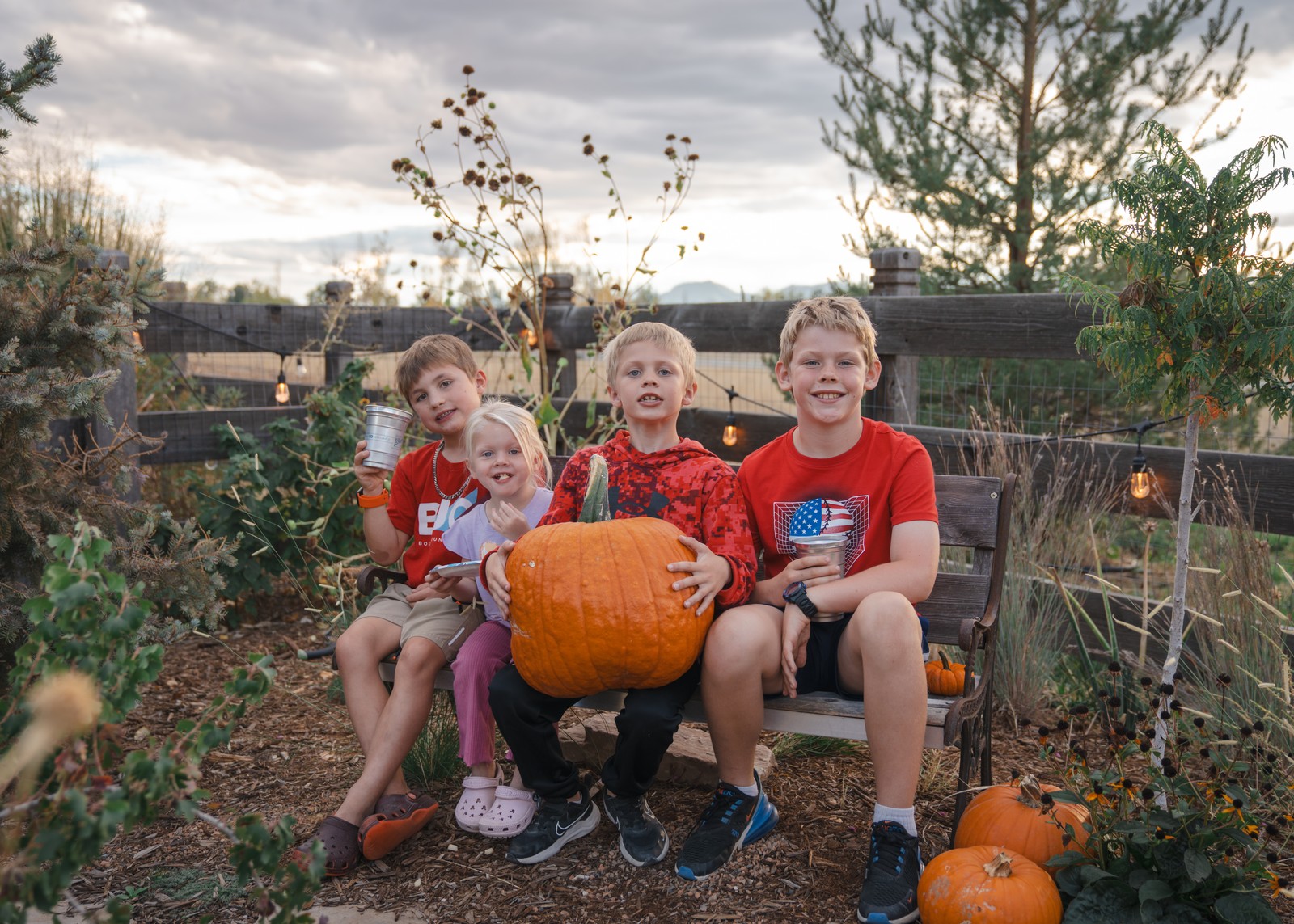 Three people sit on a garden bench behind a large pumpkin, wearing red tops, with more pumpkins on the ground.