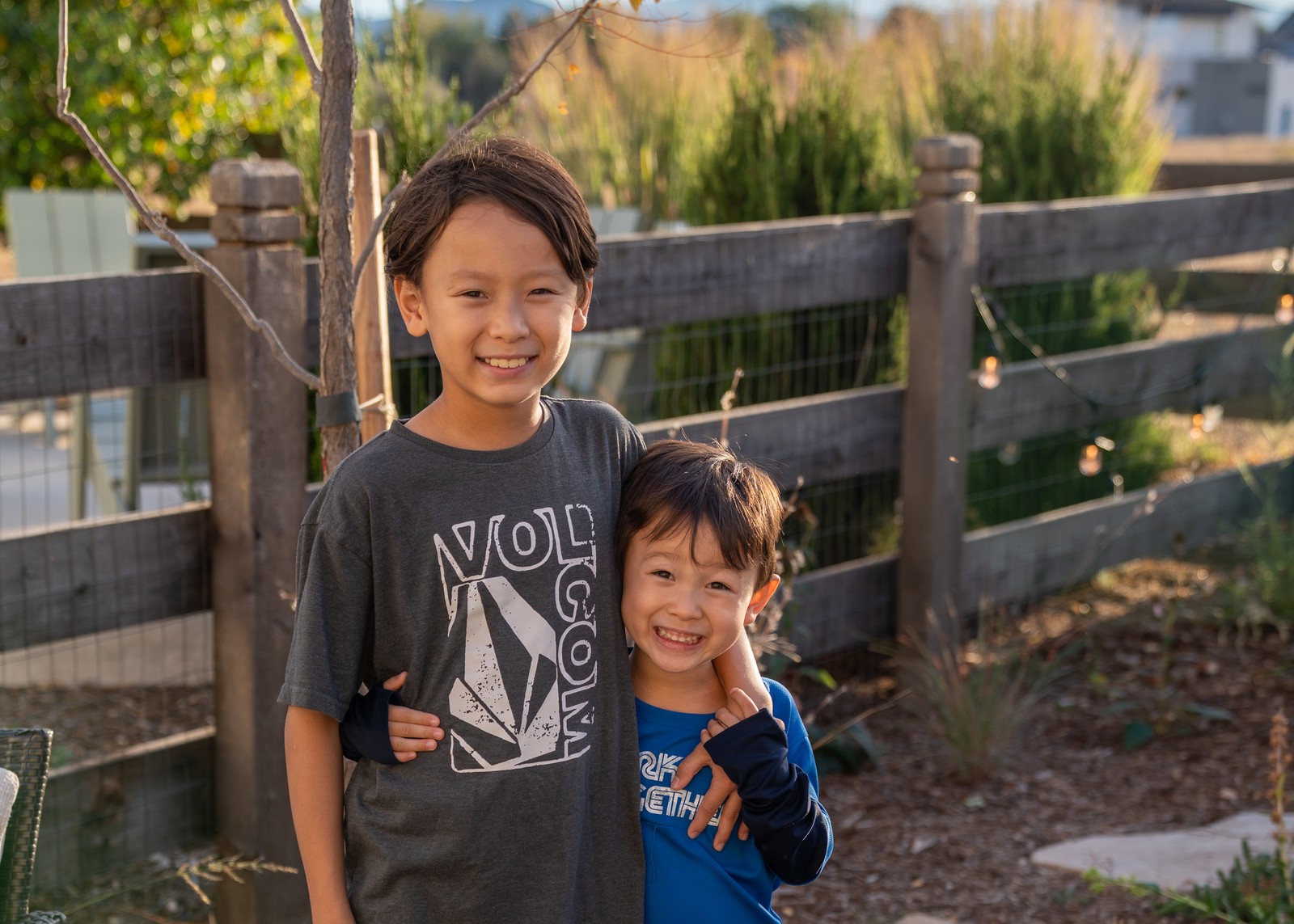 Two people smile close together in front of a wooden fence, one in a gray graphic t-shirt and one in a blue shirt.