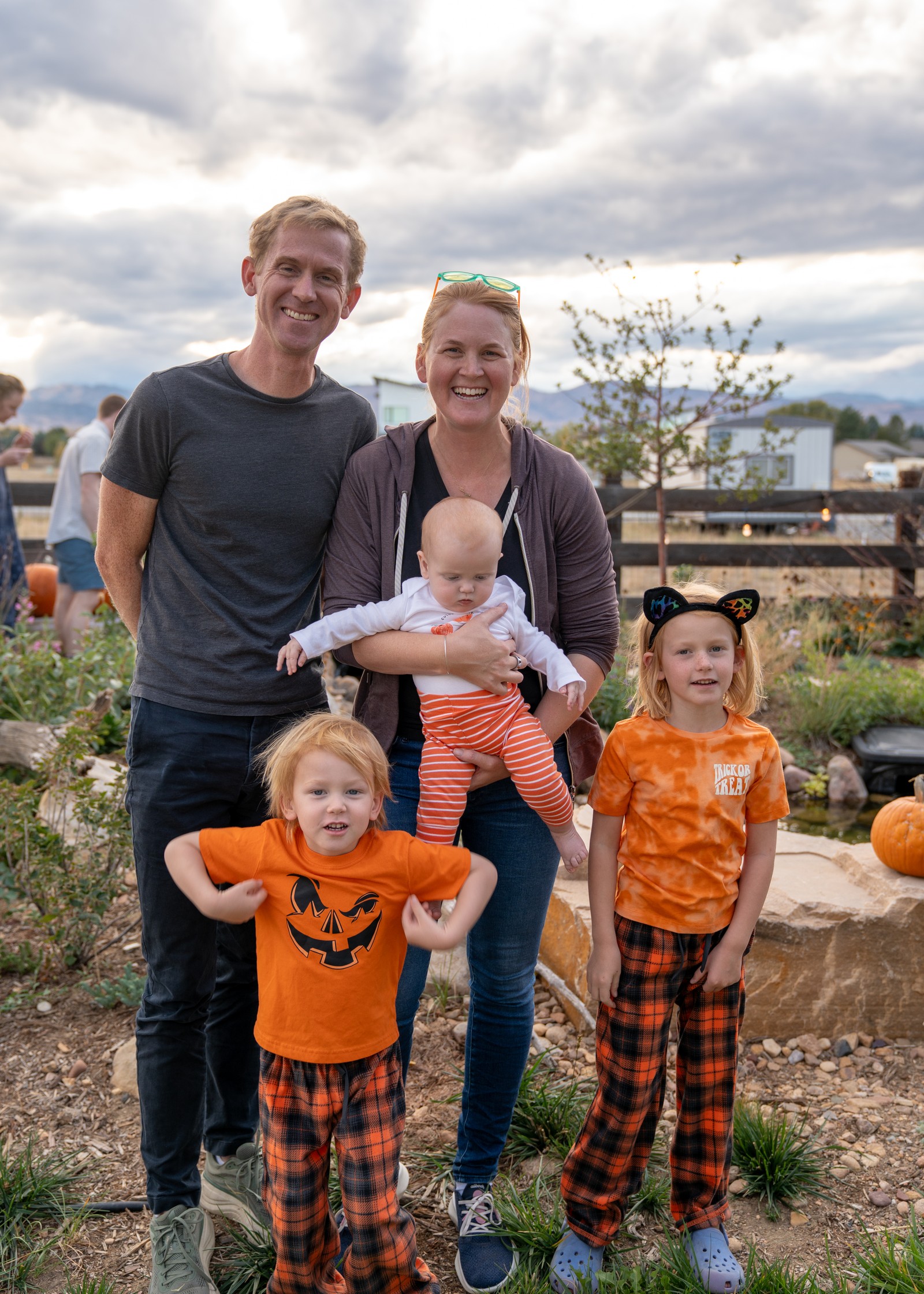 A group stands together in a yard at dusk, with two people in matching orange pumpkin shirts and plaid pants.