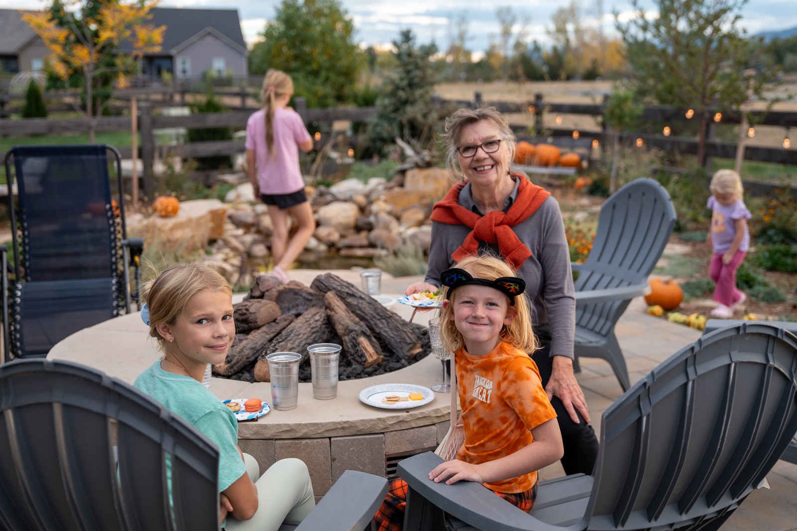 People sit around a stone fire pit table on a patio at dusk, one in an orange top and cap, with a person walking in the background.
