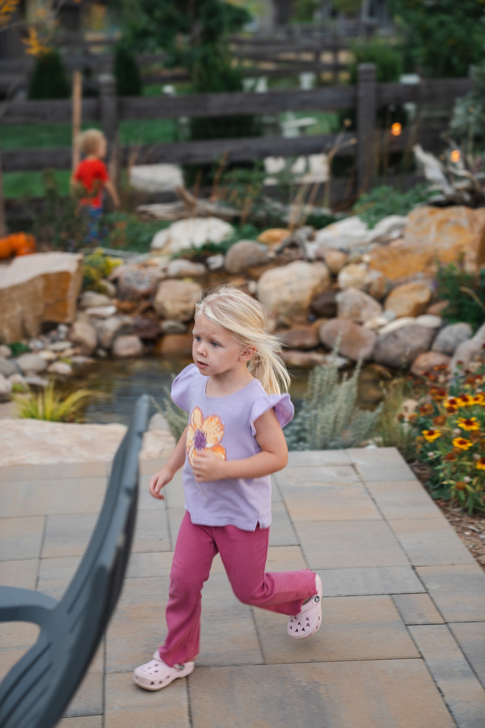 A person in a pink top and pink pants walks quickly across a paver patio beside a rock-lined pond.