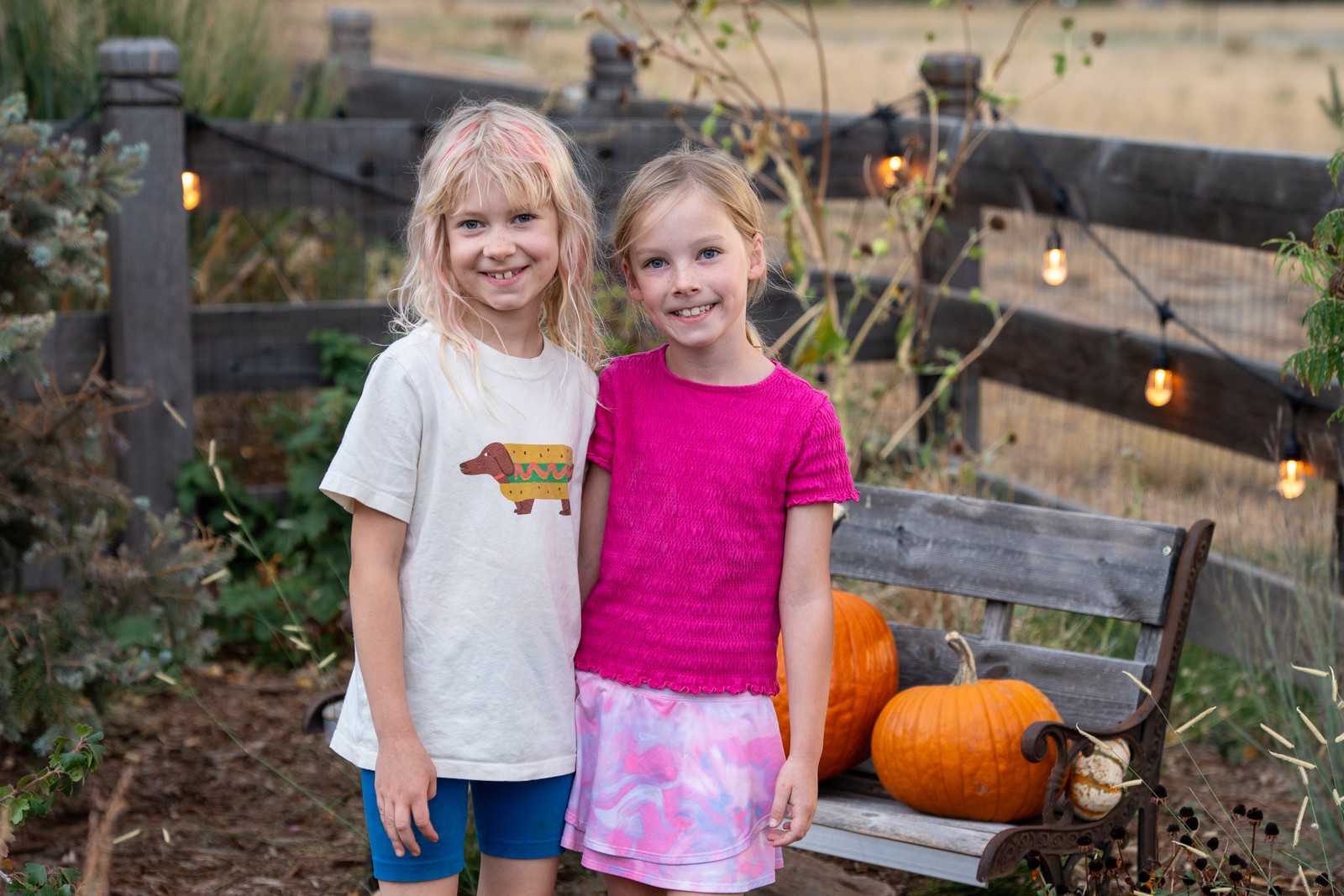 Two people stand side by side next to a wooden bench with pumpkins, one in a white t-shirt and one in a bright pink top, with string lights behind them.