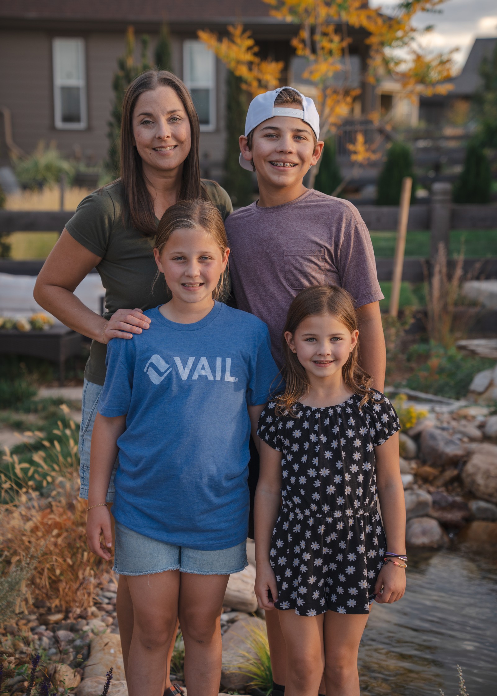 Four people pose close together beside a pond, with two in blue and dark patterned outfits and houses and trees behind them.