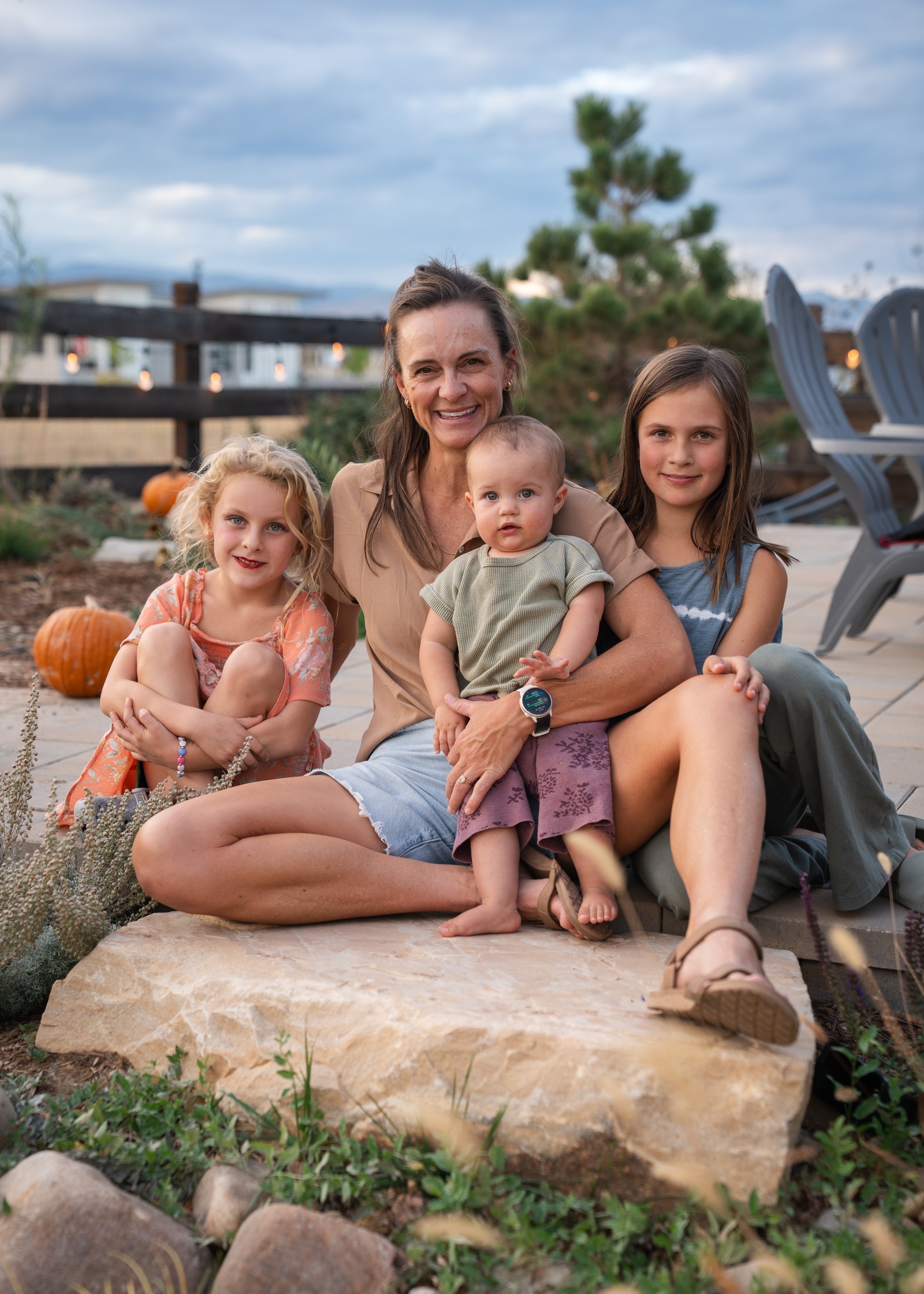 A group sits together on a flat boulder in a garden, with evergreens and a wooden fence behind them.